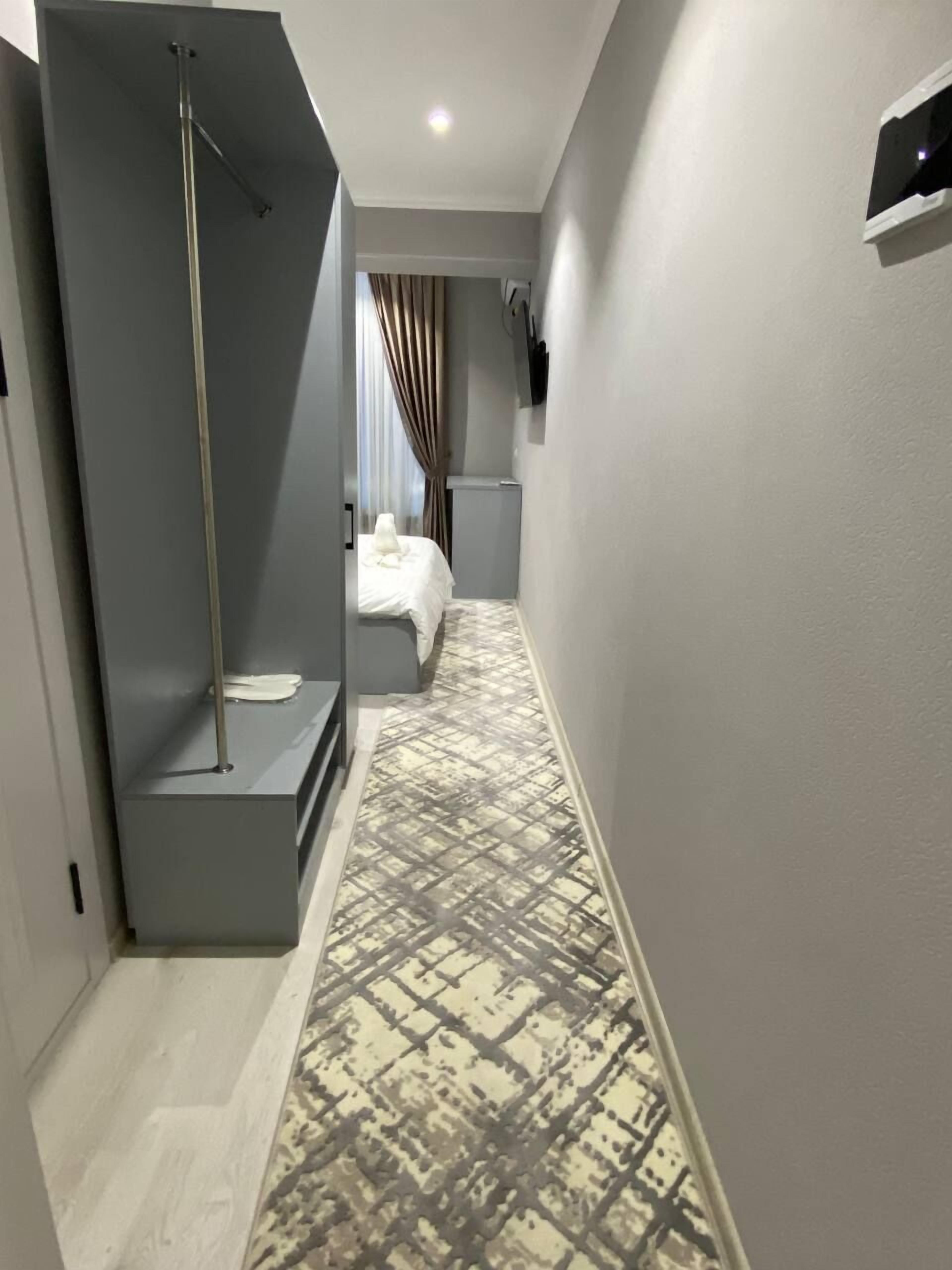 A narrow hallway with a patterned carpet, a bed with white bedding, and a shower area with a vertical pole.