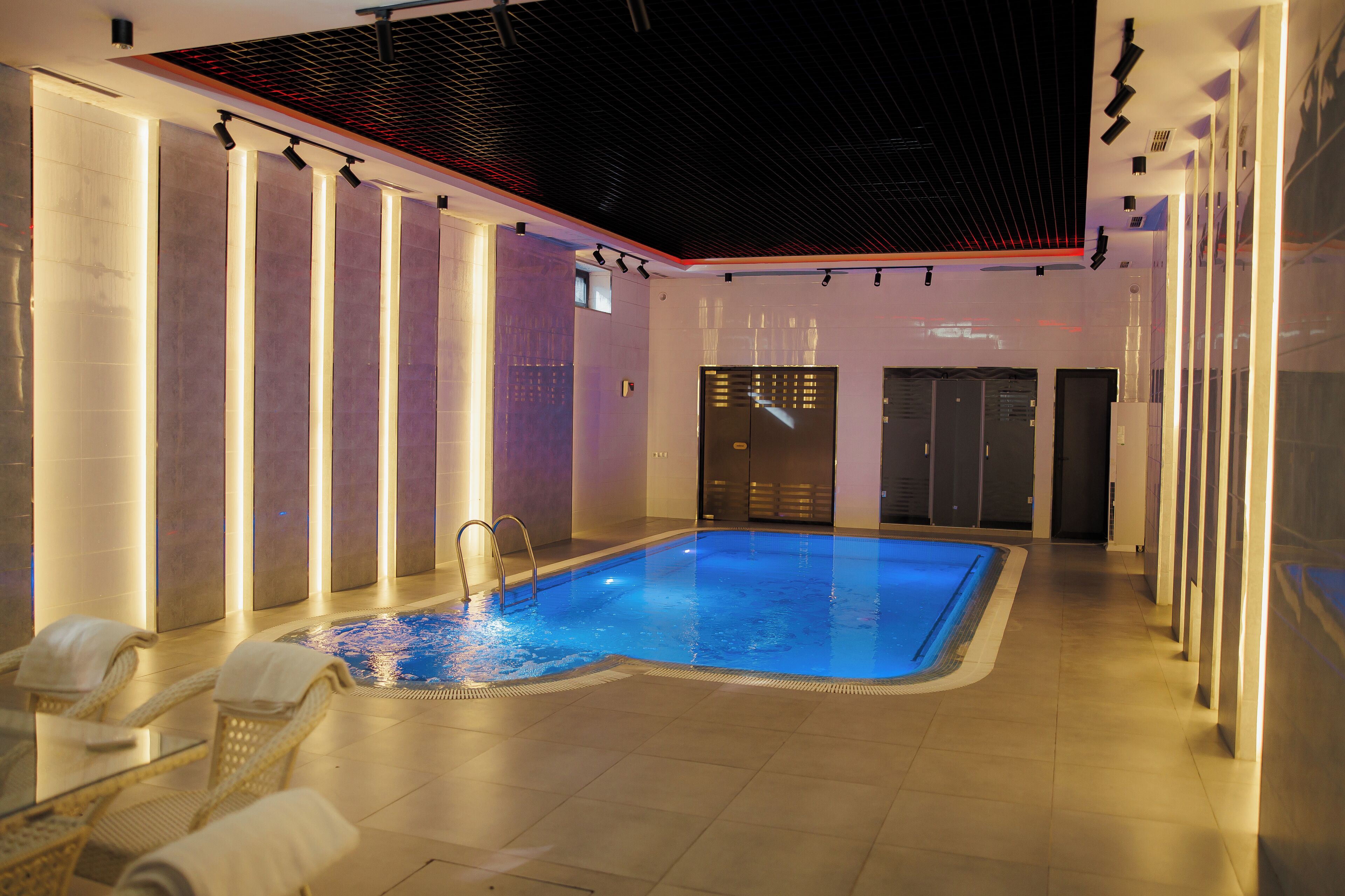 An indoor swimming pool with a tiled floor, lounge chairs with towels, and a ceiling with recessed lighting.