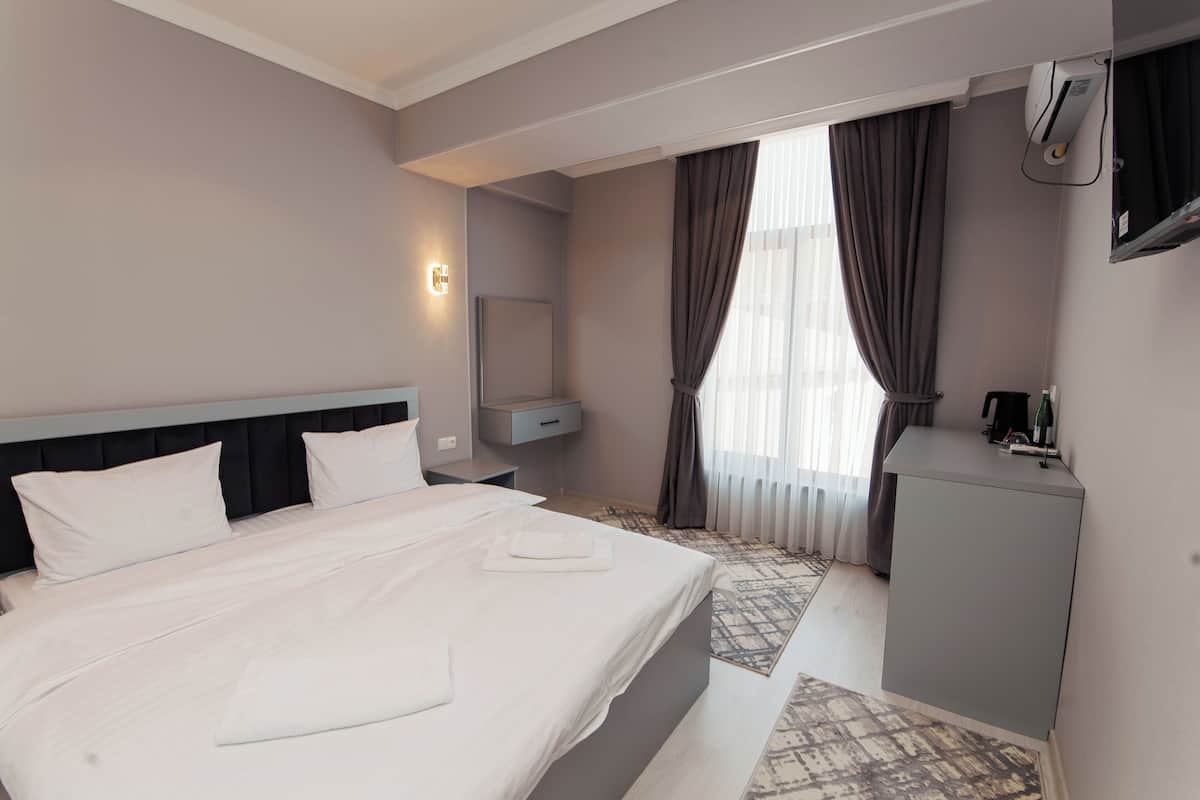 A hotel room with a large bed, a bedside table, a desk, a chair, a mirror, and a window with curtains.