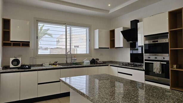 A modern kitchen with white cabinetry, granite countertops, a built-in oven, and a window with blinds. - Charming villa on the dream island of Djerba (Tunis)