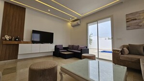 A modern living room with a flat-screen TV, comfortable seating, a glass coffee table, and a view of a swimming pool through large sliding doors. - Charming villa on the dream island of Djerba (Tunis)
