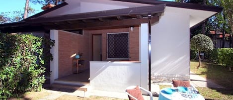 A small, covered outdoor area with a table and chairs, a window with a metal screen, and a brick wall.
