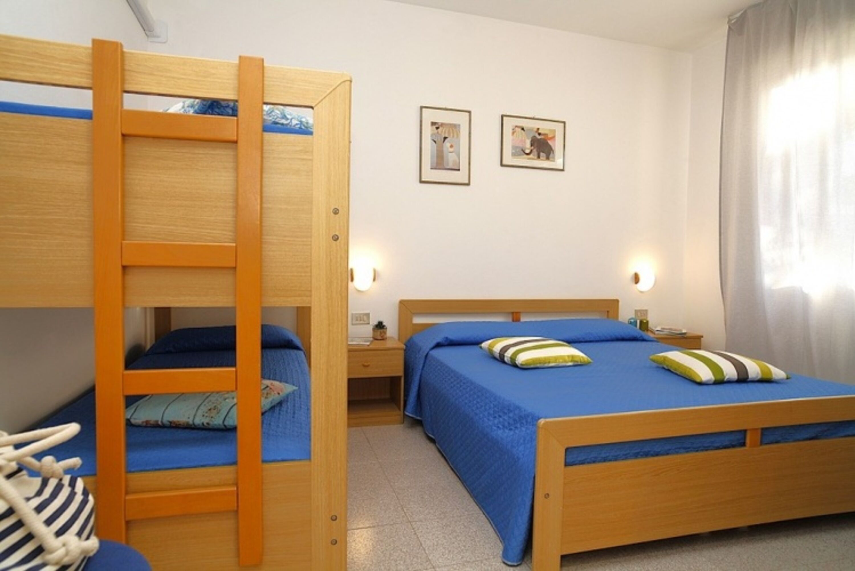 A room with bunk beds, a bed with blue bedding, bedside tables, wall-mounted lights, and framed pictures on the wall.