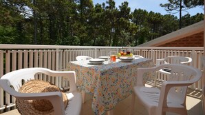 Une terrasse avec une table mise pour deux, entourée de chaises blanches, sur laquelle on trouve un panier de fruits et un verre rempli de boisson.