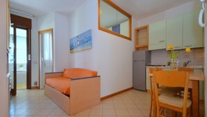 Un espacio habitable compacto con sofá cama, mesa de comedor con sillas, zona de cocina y un espejo grande.