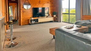 L’intérieur d’un chalet accueillant, avec un plafond en bois, une grande télévision posée sur un meuble en bois, un canapé et une table à manger.