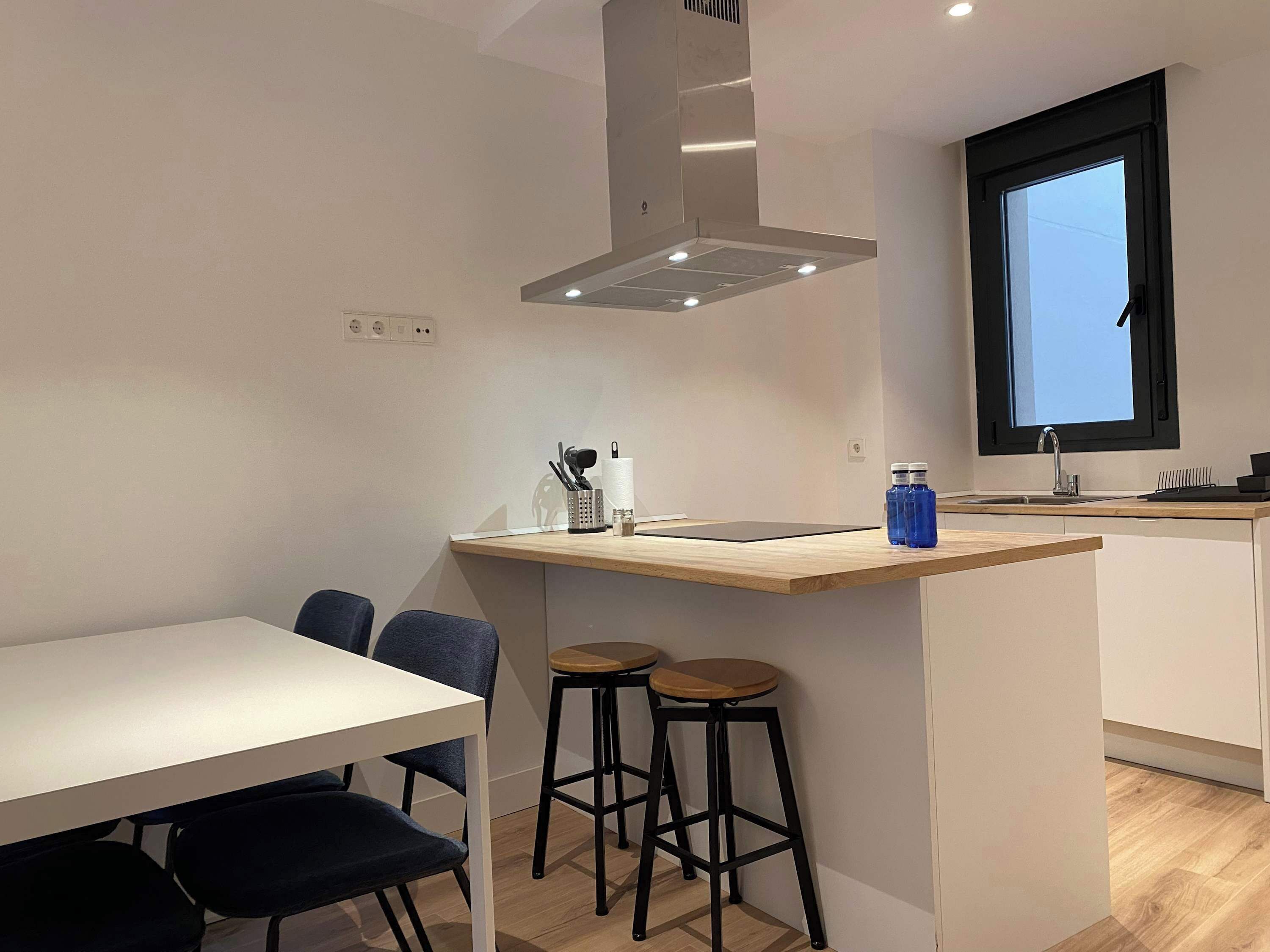 A modern kitchen with a central island, bar stools, a dining table, and a window.