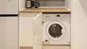 A compact kitchen with a built-in washing machine, a coffee maker, and a shelf with glassware and plates. - w* | Majestic 2BR at Palacio Real (Madrid)