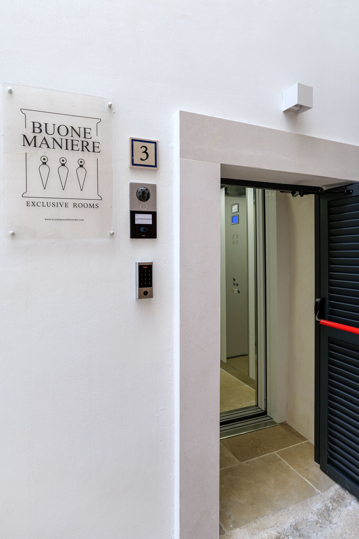 Une entrée d’ascenseur avec une enseigne indiquant « Buone Maniere, chambres de luxe » et le chiffre « 3 » au-dessus.