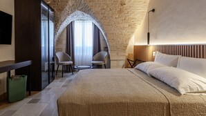 A hotel room with a stone wall, a bed with white linens, two chairs, a small table, and a TV on the wall. - buone maniere exclusive room (Martina Franca)