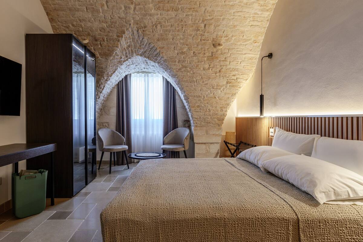 Une chambre d’hôtel avec un mur en pierre, un lit avec du linge de maison blanc, deux chaises, une petite table et un téléviseur fixé au mur.