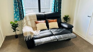 A black leather sofa with a white throw blanket and pillows, a small potted plant, and a window with blue curtains. - Your separate space in Wigram. (Christchurch)