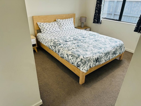 A bedroom with a wooden bed, floral bedding, a nightstand, a lamp, and a window with curtains. - Your separate space in Wigram. (Christchurch)