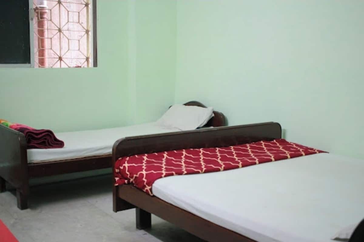 A room with two single beds, each with a wooden frame and a mattress. There is a window with a metal grid pattern, and the walls are painted in a light color.