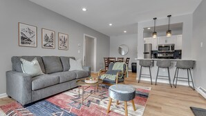 A modern living room with a grey sofa, a patterned rug, and a dining area with bar stools.