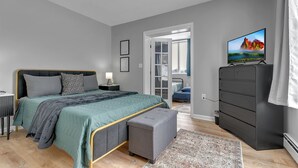 A modern bedroom with a bed, bedside table, TV, dresser, and a view of a cityscape through a door.