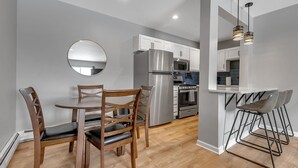 A modern kitchen with a dining area, featuring a round mirror, wooden chairs, a stainless steel refrigerator, a microwave, and a bar area with stools.