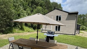 A wooden table with four chairs and a large umbrella, set on a gravel surface with a grill and a house in the background. - Ellicottville 2 miles to Holiday Valley Condo 4 (Great Valley)