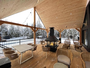 Una terraza de madera con chimenea, mobiliario de mimbre y vistas a árboles cubiertos de nieve.