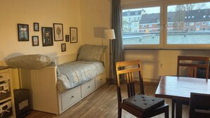 Una habitación pequeña con cama, una silla, una mesa y ventana con vista a una zona residencial.