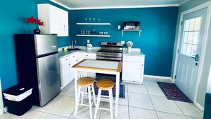 Een compacte keuken met witte kastjes, een roestvrijstalen koelkast, een magnetron en een fornuis. Er staan twee krukken bij een klein tafeltje, en een prullenbak is zichtbaar.