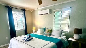 Een hotelkamer met een bed, twee ramen met zonwering, een airconditioning, een bedlampje en een kleine tafel met lamp.