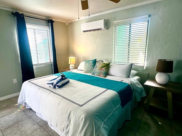 A hotel room with a bed, two windows with blinds, an air conditioning unit, a bedside lamp, and a small table with a lamp.