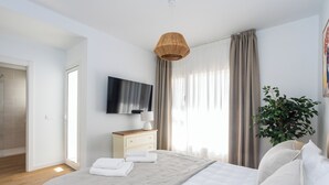Moderni hotellihuone, jossa on suuri sänky, taulutelevisio, yöpöytä lampun kanssa ja ruukkukasvi.