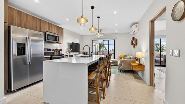 A modern kitchen with stainless steel appliances, a central island with bar stools, pendant lighting, and a view to a living area with a sofa and lamp.