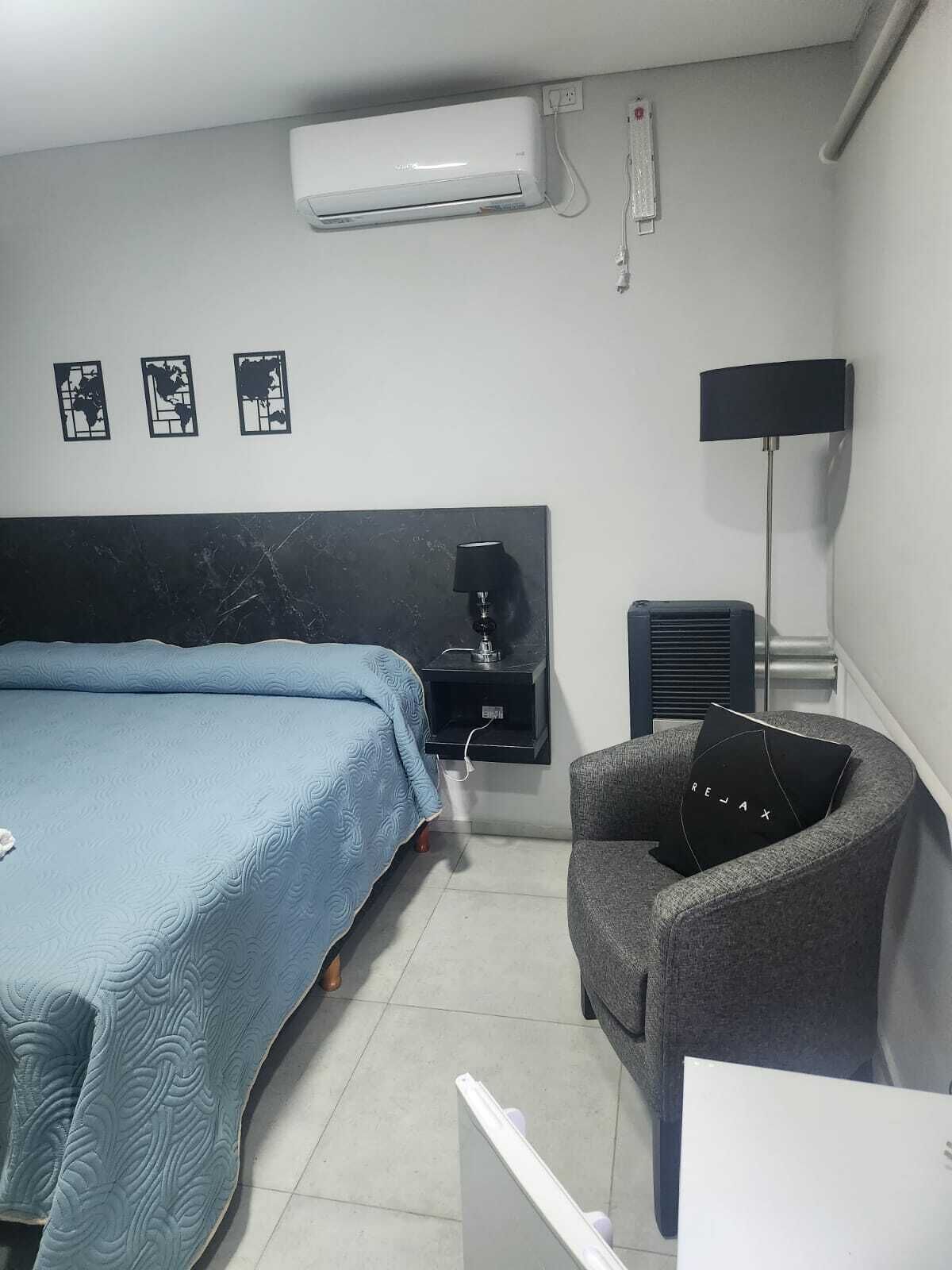 A hotel room with a bed, a desk, a chair, a lamp, and an air conditioning unit.