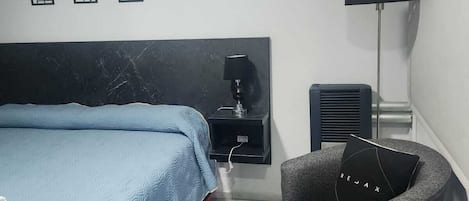 A hotel room with a bed, a desk, a chair, a lamp, and an air conditioning unit.