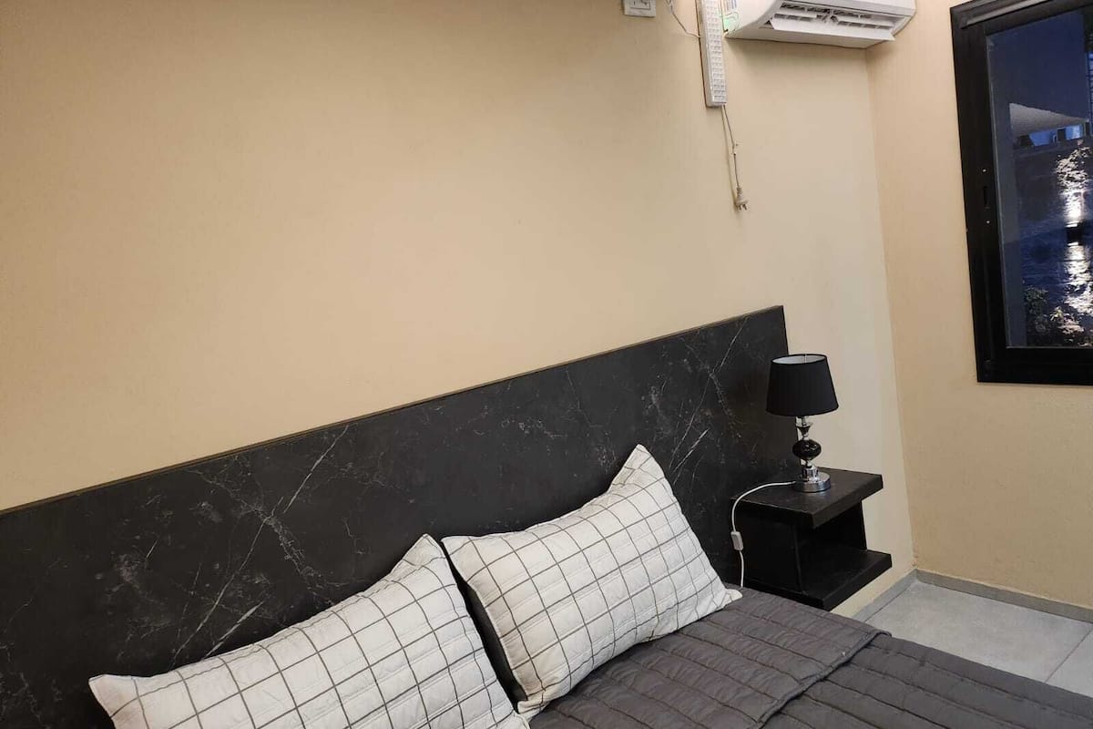 A hotel room with a bed, a nightstand, a lamp, an air conditioning unit, and a window with a night view.