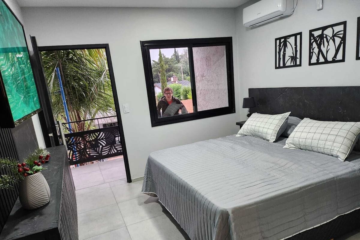 A bedroom with a bed, a desk, a chair, a potted plant, and a window with a view of greenery.