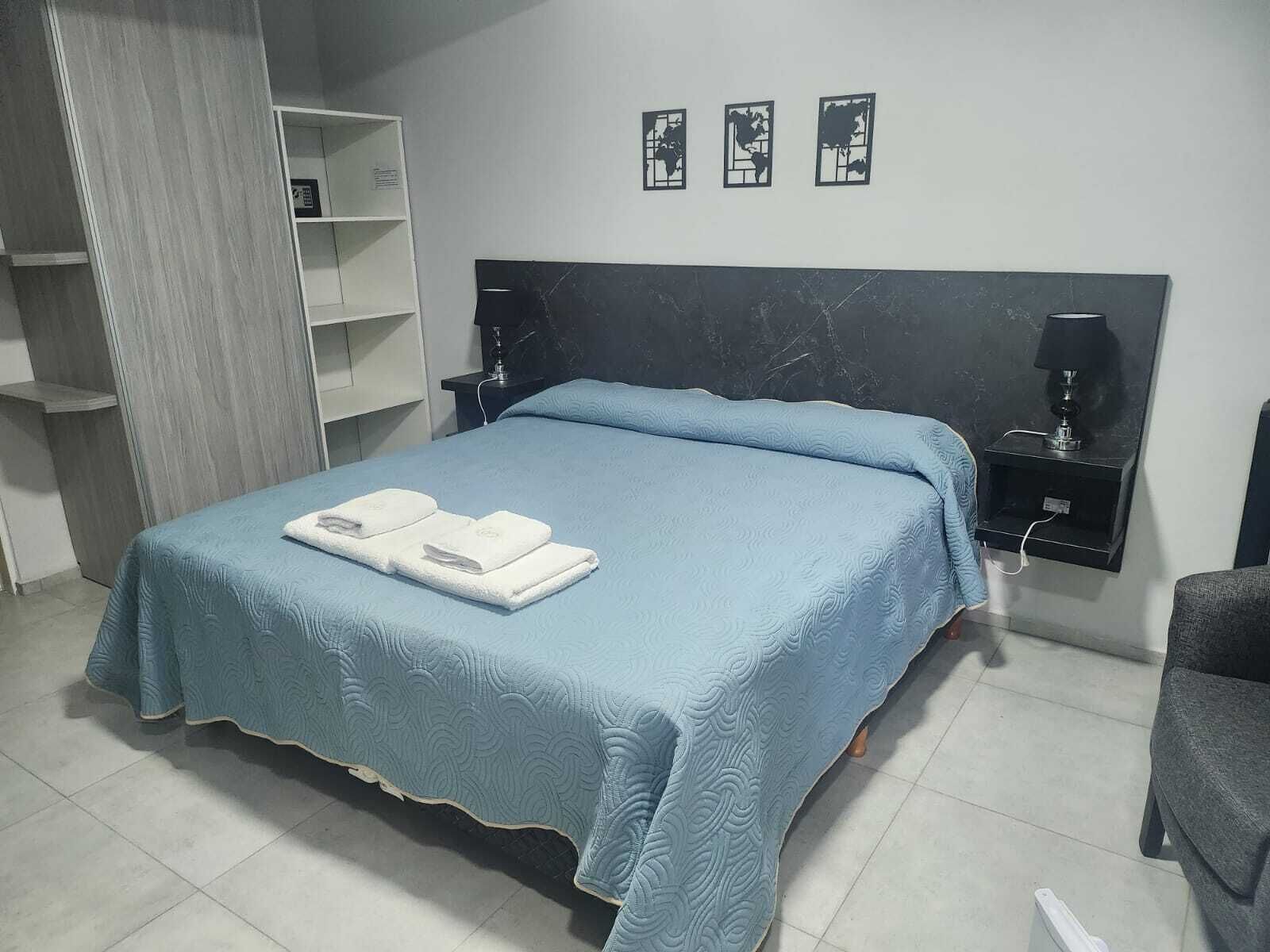A neatly made bed with a blue bedspread and two folded white towels, a dark headboard, a small shelf with a lamp, and three framed pictures on the wall.
