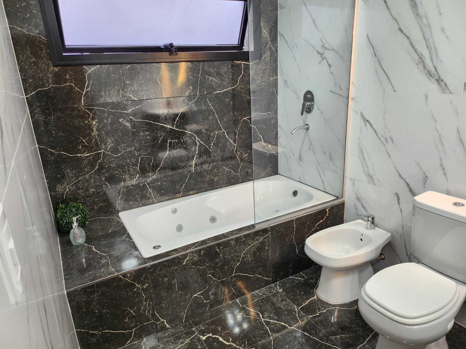 Une salle de bain moderne dotée d’une baignoire, d’un bidet et de toilettes, avec des murs en marbre foncé et un grand miroir.