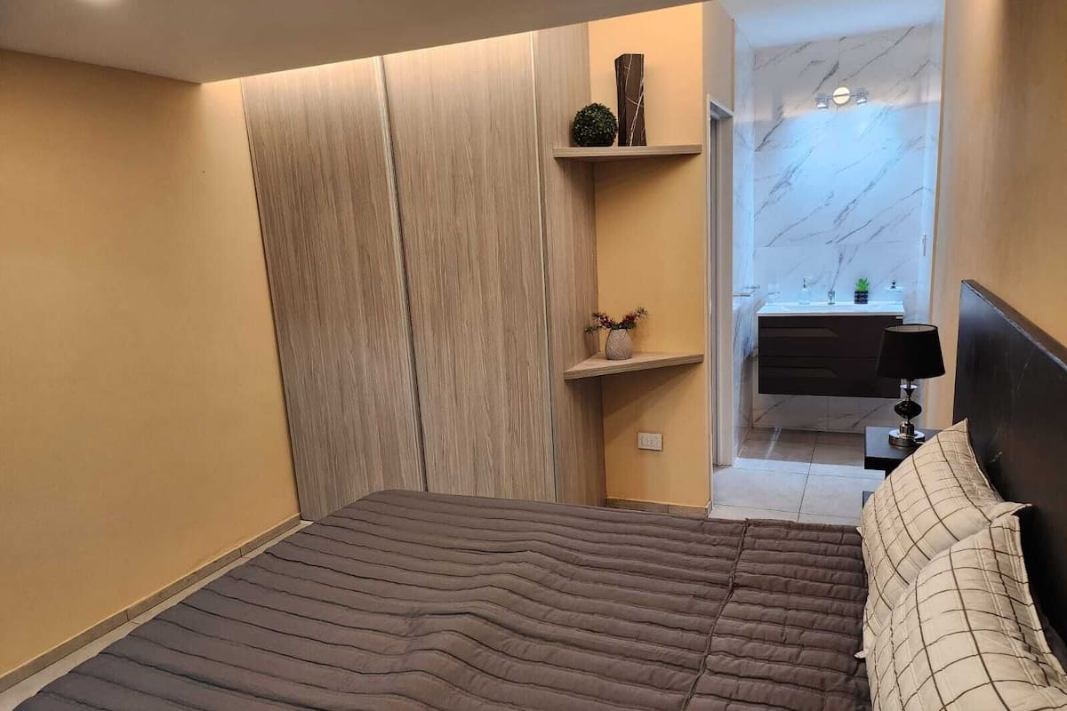 A modern bedroom with a large bed, a bedside table with a lamp, and a small shelf with decorative items.