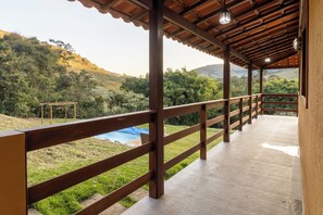 Área externa coberta com deck de madeira, grades de metal e vista para um morro com árvores e uma lona azul.