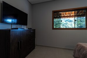Quarto com televisão de tela plana na parede, janela com vista para árvores e um armário de madeira escura.