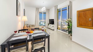 A modern living room with a dining area, large windows with city views, a flat-screen TV, and a wooden cabinet. - Stylish 1BR Retreat in Dubai | Lake Views, Pool, Gym & Prime City Location (Dubai Production City)