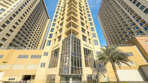 A high-rise building with a central glass facade and multiple balconies. - Stylish 1BR Retreat in Dubai | Lake Views, Pool, Gym & Prime City Location (Dubai Production City)