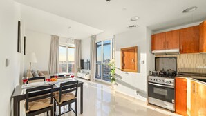 A modern kitchen with a stove, oven, and wooden cabinets. There is a dining area with a table and chairs, and a large window offering a city view. - Stylish 1BR Retreat in Dubai | Lake Views, Pool, Gym & Prime City Location (Dubai Production City)