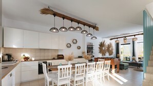 Une cuisine moderne avec des meubles blancs, un îlot central doté de sièges, un éclairage suspendu et une vue sur l’espace de vie.