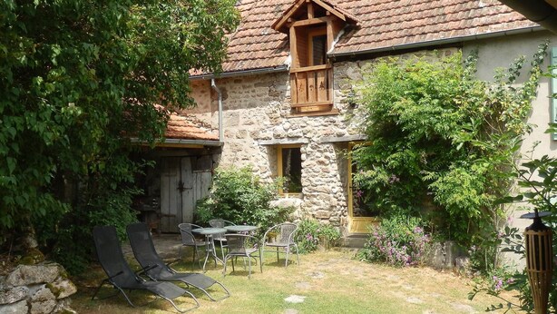 Ein Steinhaus mit Ziegeldach, einem Holster und einem kleinen Garten mit Rasen, einer hölzernen Bank und einem Tisch mit Stühlen.