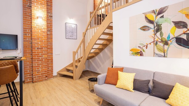 A modern living room with a wooden staircase, a grey sofa with yellow cushions, a brick accent wall, and a framed cityscape artwork. - Soulscape Apartment CityCenter - compact & luxurious loft apartment in the city center (Zwickau)