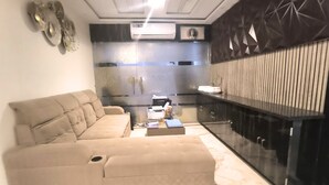 A modern living room with a beige sectional sofa and a small coffee table. A kitchen with a black countertop and stainless steel appliances is visible in the background. - Hotel Devvanshi Vila (Varanasi)