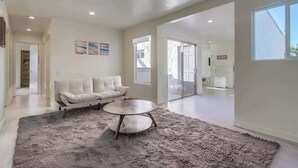 A spacious living room with a white sofa, a round coffee table, and a shaggy rug. There are two paintings on the wall and a sliding glass door leading to another room. - Ocean views, steps to beach, family, dining, shops (San Diego)