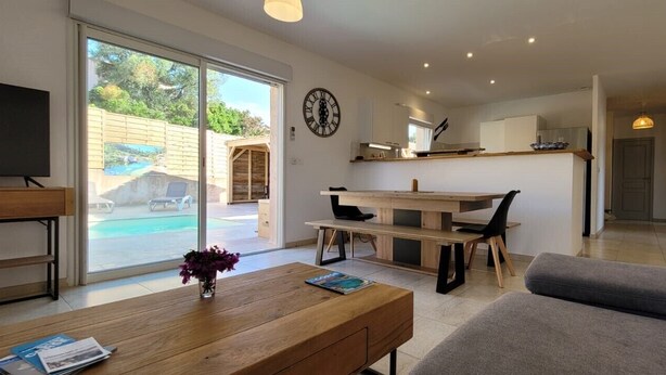 A modern living room with a wooden coffee table, a grey sofa, and a dining area with a wooden table and chairs. A TV is mounted on the wall, and a large window offers a view of a pool and outdoor area. - Villa Porto Vecchio, near beach, 8/10 pers (sainte lucie de Porto vecchio)