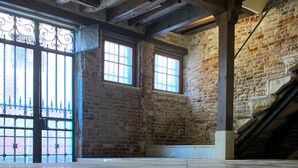 A spacious room with exposed brick walls, large windows, and a tiled floor.