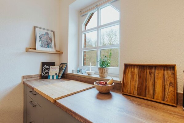 A kitchen with a wooden countertop, a bowl of fruit, a framed picture, and a window with a view of trees.