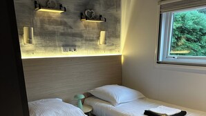Een compacte hotelkamer met twee eenpersoonsbedden, een klein nachtkastje, een wandplank met decoratieve objecten, en een raam met uitzicht op groen.